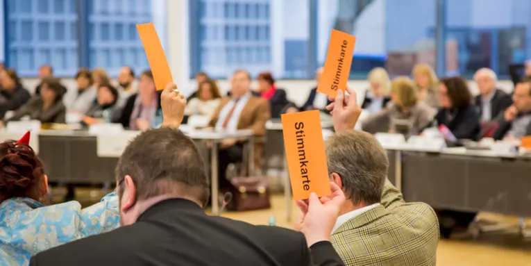 Mitglieder der Regionsversammlung heben orangefarbene Pappkarten mit dem schwarzen Text "Stimmkarte" in die Luft.