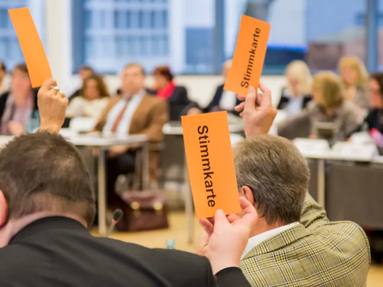 Mitglieder der Regionsversammlung heben orangefarbene Pappkarten mit dem schwarzen Text "Stimmkarte" in die Luft.