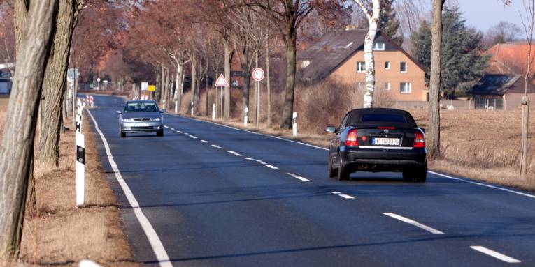 Die Fahrbahn einer Kreisstraße wird aus beiden Richtungen von jeweils einem Auto befahren, an der rechten Seite sind ein Vehrkehrsschild "Geschwindigkeitsbegrenzung 70 km/h", "Fahrbahneinengung" und ein gelbes Ortsschild zu sehen, im Hintergrund beginnt eine Wohnsiedlung.