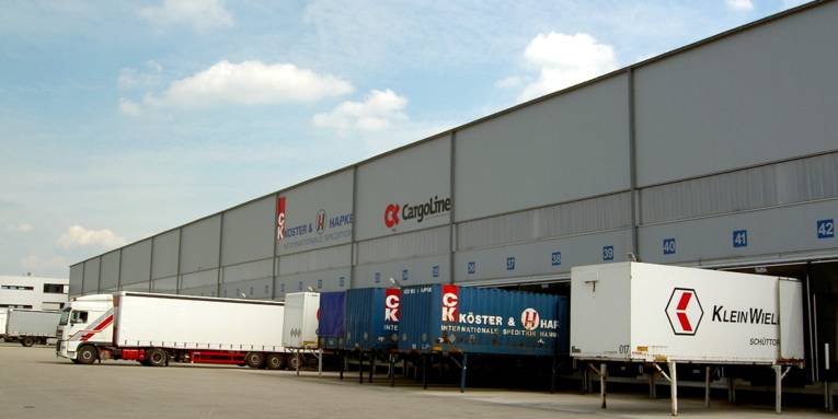 Vor einem großen Lagergebäude stehen mehrere Lkw nebeneinander an den Laderampen.