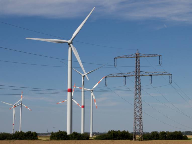 Die linke Bildhälfte wird von sechs Windkraftanlagen des Windparks zwischen Pattensen und Sarstedt dominiert, die rechte Hälfte beherrscht ein Hochspannungsmast. Im Hintergrund zeichnet sich ein Getreidefeld vor blauem Himmel ab.