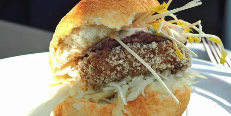 Zwischen zwei hellen, knusprigen Brötchenhälften sind Krautsalat, eine vegetarische Bulette, Rotkrautdip, Knoblauchsoße und Sprossen übereinander gestapelt. Der vegetarische Burger liegt auf einem Teller und wird von links von Sonnenlicht beschienen. Im Hintergrund liegt eine Gabel.