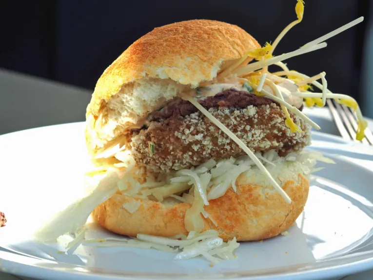 Zwischen zwei hellen, knusprigen Brötchenhälften sind Krautsalat, eine vegetarische Bulette, Rotkrautdip, Knoblauchsoße und Sprossen übereinander gestapelt. Der vegetarische Burger liegt auf einem Teller und wird von links von Sonnenlicht beschienen. Im Hintergrund liegt eine Gabel.