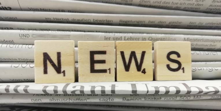 Vier Holzwürfel mit Buchstaben drauf, die nebeneinander aufgestellt das Wort "News" ergeben. Dahinter ist ein Stapel Zeitungen zu sehen.