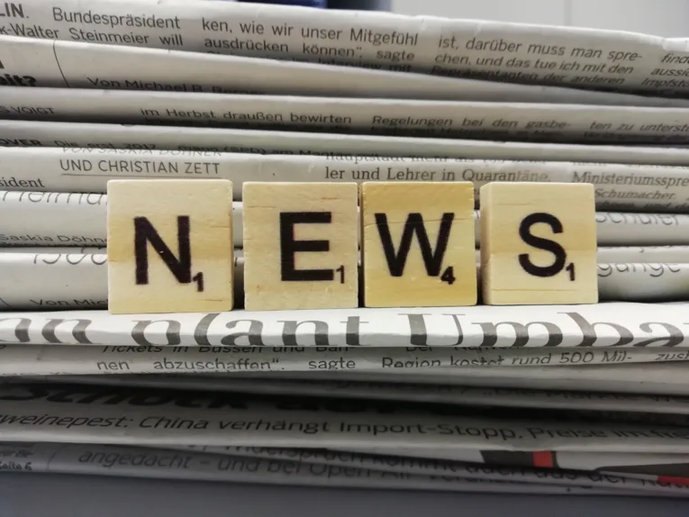 Vier Holzwürfel mit Buchstaben drauf, die nebeneinander aufgestellt das Wort "News" ergeben. Dahinter ist ein Stapel Zeitungen zu sehen.