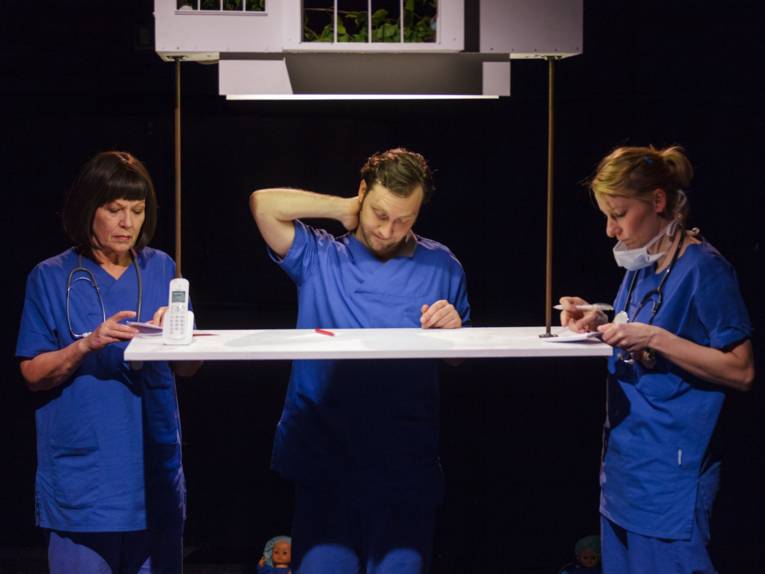 Drei Ärzte in blauen Kitteln stehen um einen erhöhten Tisch. Sie haben die Augen gesenkt und sind in das Lesen von Unterlagen vertieft.