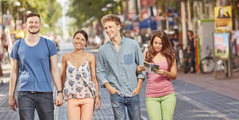 Vier junge Erwachsene spazieren lachend durch eine belebte Fußgängerzone, eine von ihnen liest eine Stadtkarte.