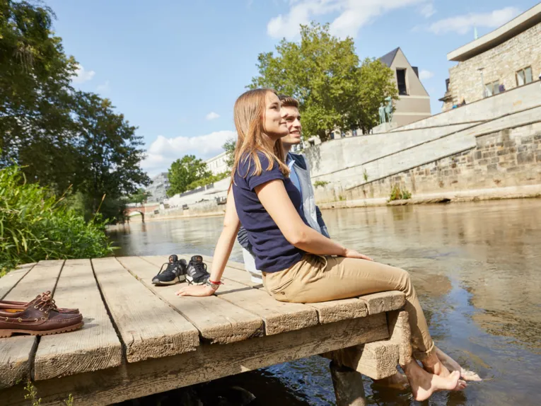 Ein junges Paar sitzt auf einem Holzsteg am Hohen Ufer und lässt die Füße ins Wasser hängen. Schuhe liegen daneben, Stadtmauer im Hintergrund.