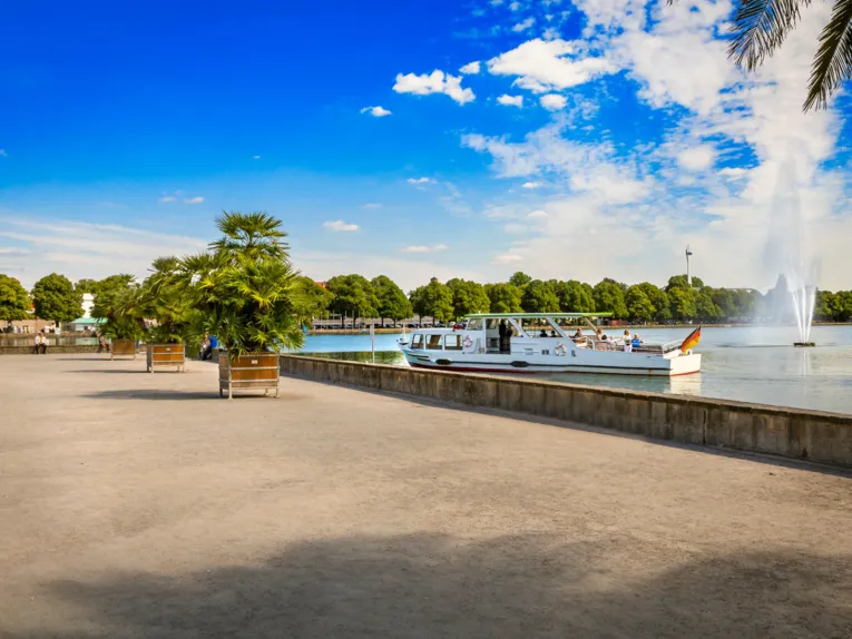 Ein Ausflugschiff mit Passagieren fährt auf dem Maschsee mit großer Fontäne, während Palmen in Kübeln die Uferpromenade säumen.