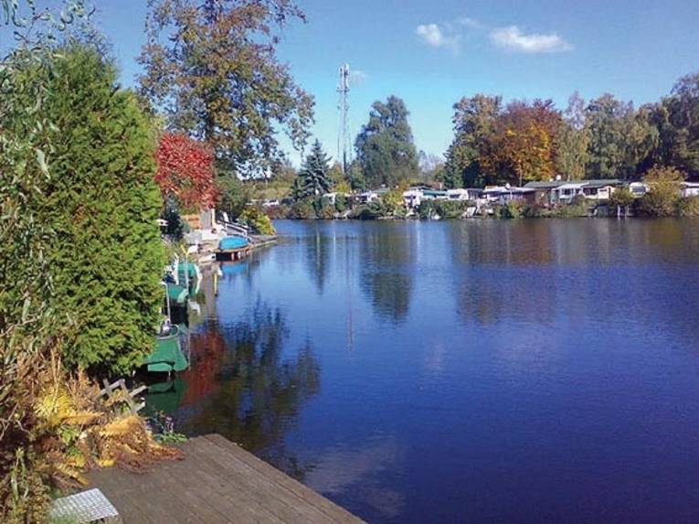 Ein See, gesäumt von herbstlich gefärbten Bäumen und einigen Wohnmobilen im Hintergrund. 