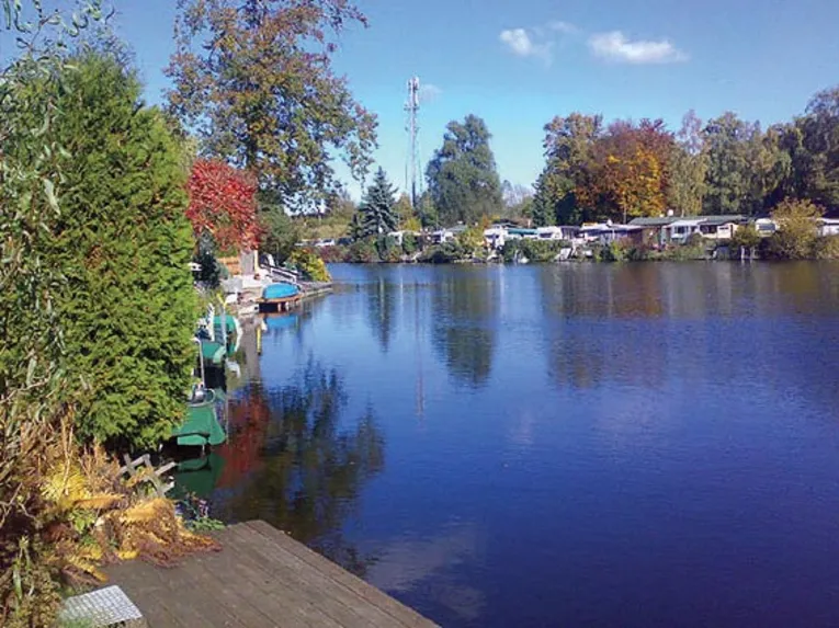 Ein See, gesäumt von herbstlich gefärbten Bäumen und einigen Wohnmobilen im Hintergrund. 