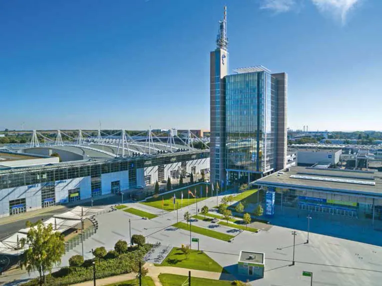 Luftaufnahme des Messegeländes Hannover mit Glasbürogebäude, Funkturm, Messehallen und weitem Vorplatz bei sonnigem Wetter.