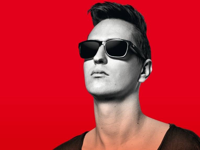 Junger Mann in schwarz-weiß mit schwarzer Sonnenbrille vor einem roten Hintergrund.