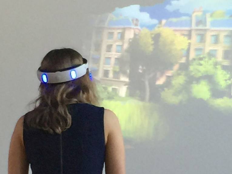 Junge Frau mit leuchtendem Virtual Reality Stirnband steht vor einer Wand, auf der ein Bild eines Gebäudes erzeugt wird.