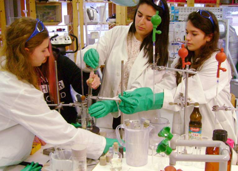 Drei Schülerinnen in weißen Kitteln und grünen Versuchshandschuhen hantieren in einer Laborumgebung mit Reagenzgläsern und Proben
