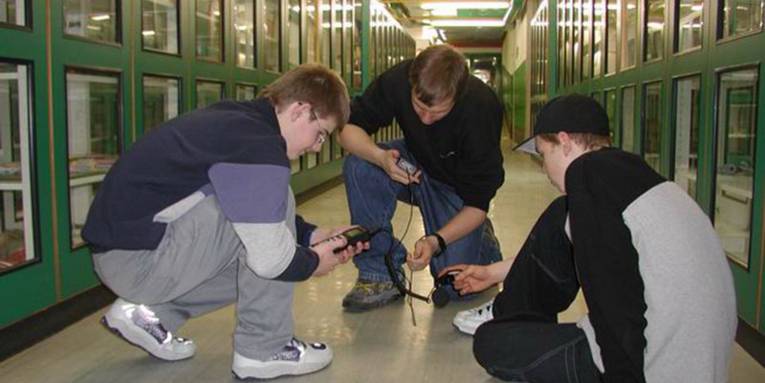 Drei Schüler knien auf einem hell erleuchteten Schulkorridor und messen die Lichtstärke
