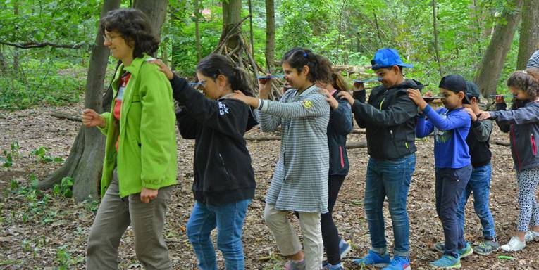Mehrere Kinder gehen mit einer Betreuerin durch einen Wald und halten sich jeweils an der vorderen Person mit einer Hand an deren Schulter fest.