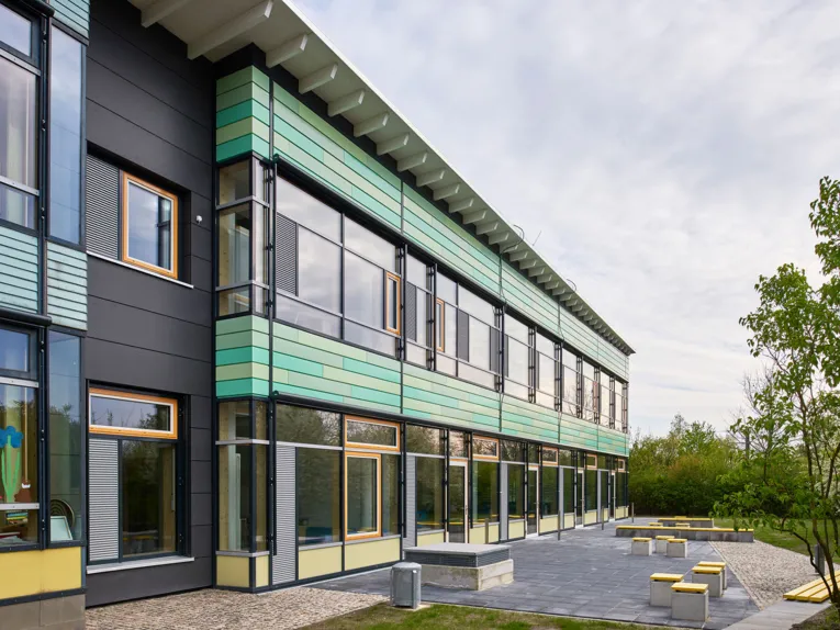 Die Außenansicht der Grundschule An der Feldbuschwende zeigt einen zweistöckigen Flachbau mit großen Fensterfronten und grüner Verkleidung, davor einen Pausenbereich mit Sitzelementen.