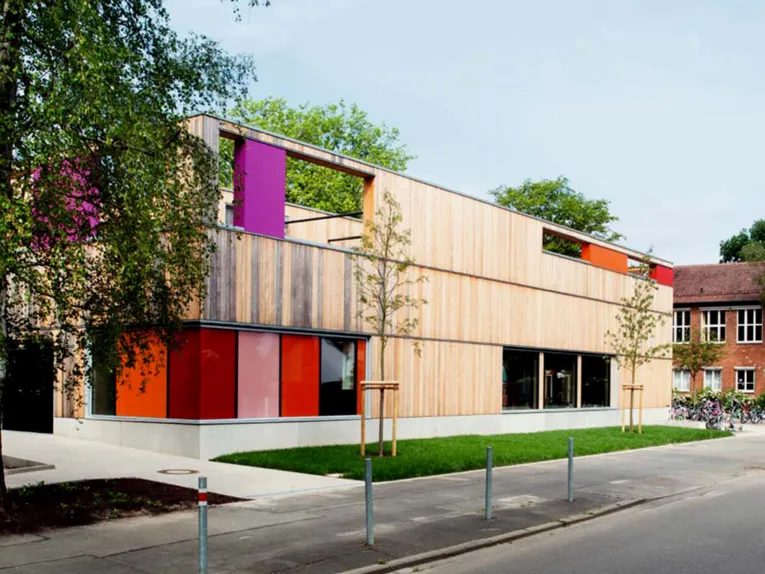 Die neugebaute Mensa der Grundschule Entenfang ist ein holzverkleideter Kubus mit barrierefreiem Zugang und bunten Tür- und Fensterelementen in orange, fliederfarben und rot.