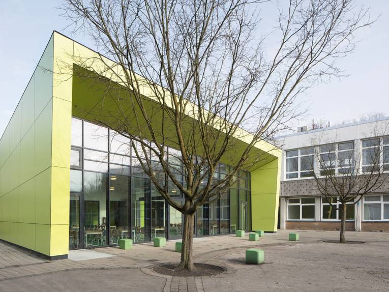 Der asymmetrisch geformte Neubau der Mensa ist an den Seiten mit hellgrünen Elementen verschalt und verfügt über eine durchgehende Fensterfront. Das Gebäude befindet sich neben dem zweigeschössigen Schulbau.