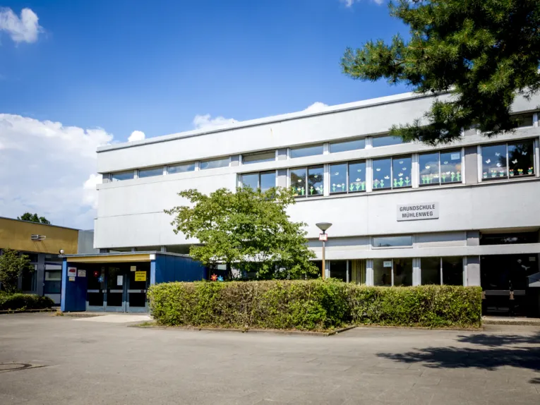 Eingang zu einem zweigeschossigen Schulgebäude, an dessen Fenster im Obergeschoss selbstgemachte Papierblumen befestigt sind. Darunter befindet sich das Schild "Grundschule Mühlenweg".