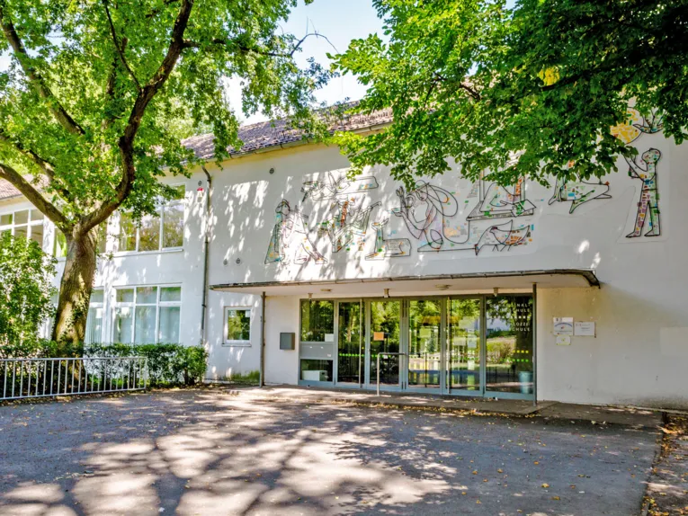 Eingang zur Pestalozzi Grundschule - ein zweistöckiges Gebäude, über dessen Eingangsdach bunte Figuren im 60er-Jahre-Stil zu sehen sind.