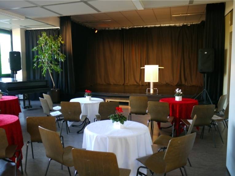 Veranstaltungssaal im Kulturtreff Plantage. Im hinteren Teil des Bildes ist die Bühne mit einem Podium zu sehen. Vor der Bühne stehen mehrere runde Tische an denen Stühle stehen. Auf den Tischen liegt abwechselnd eine rote oder eine weiße Tischdecke. Die roten Tischdecken sehen aus, als wären sie aus Samt. Auf den Tischen steht jeweils ein Blumentopf mit Alpenpfeilchen im gegengesetzter Farbe zu den Tischdecken. Dieses Aufnahme ist vor der Veranstaltung aufgenommen worden, da noch keine BesucherInnen anwesend sind.