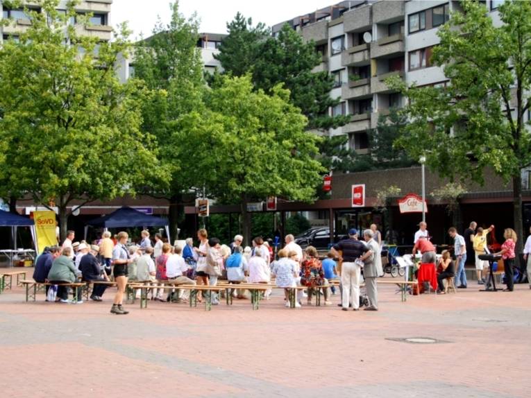 Auf dem Bild ist ein Marktplatz zu sehen, auf dem ein Fest gefeiert wird. Viele Menschen sitzen auf Bänken oder gehen über dem Marktplatz.