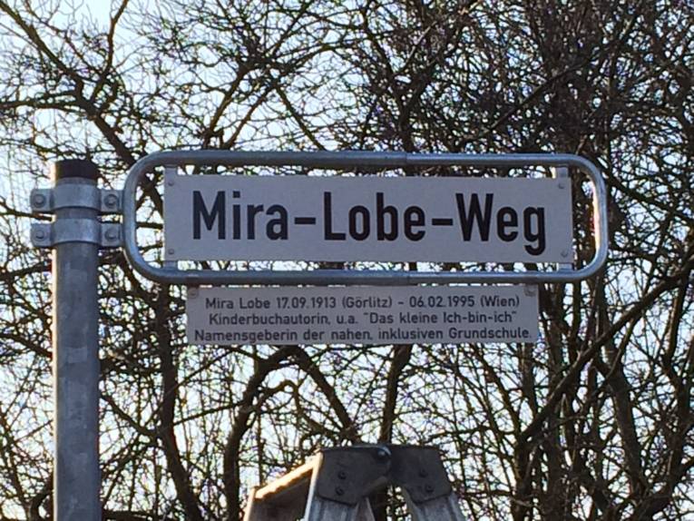 Straßenschild mit der Bezeichnung Mira-Lobe-Weg. Darunter befindet sich eine Legendentafel mit einer Kurzbeschreibung zu Mira Lobe.
