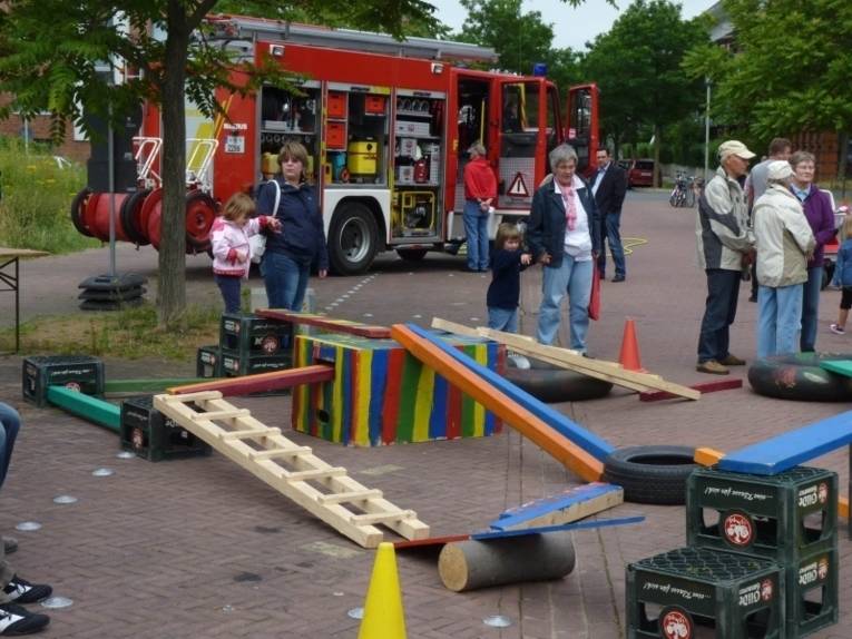 Im Vordergrund ein improvisierter Hindernis-Parcours für Kinder aus Bierkisten und bemalten Holzelementen; im Hintergrund begutachtet ein interessierter Besucher das Innere eines Feuerwehrfahrzeuges.