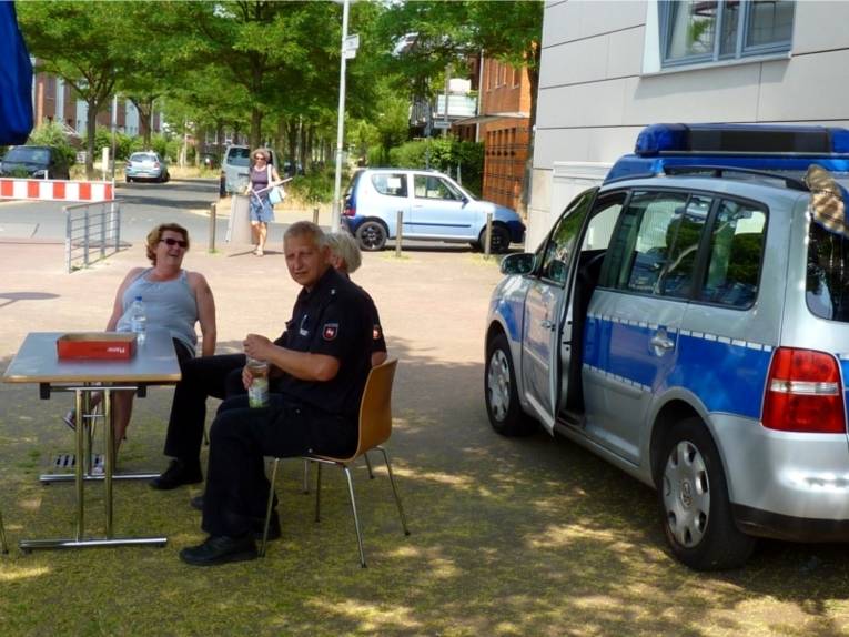 Auf dem Bild ist ein Stand mit der Polizei zu sehen. An einem Tisch sitzt ein Polizeibeamter mit zwei weiteren Damen. Hinter den drei Personen steht ein Polizeiauto.