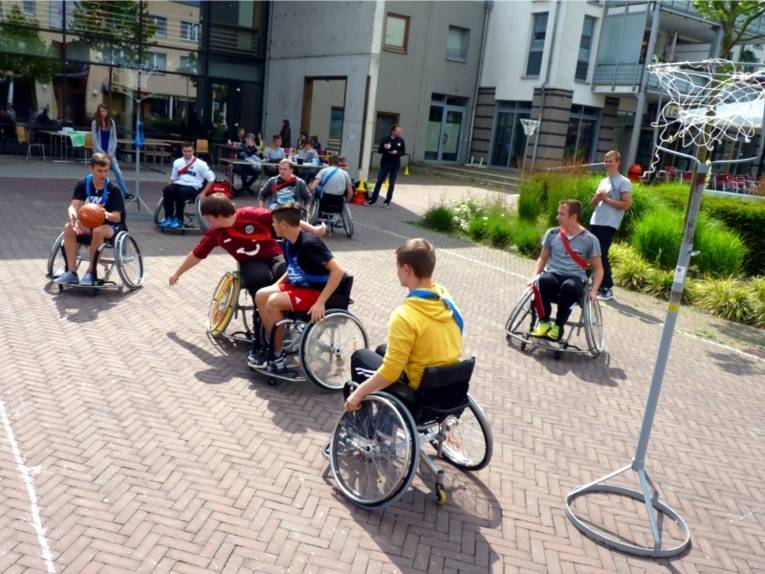 Auf dem Bild sind acht junge Rollstuhlfahrer zu sehen, die auf einem extra hergerichteten Platz mit zwei mobilen Basketballkörben Basketball spielen. 