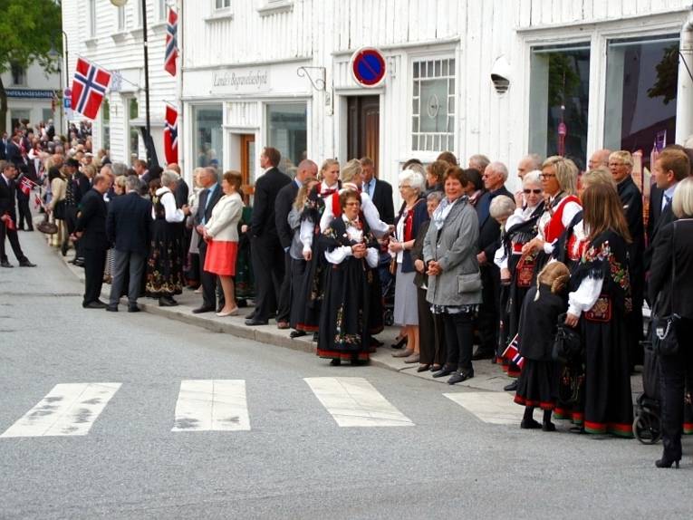Die Zuschauerinnen und Zuschauer stehen am Straßenrand und schauen sich die Parade an. Sie lächeln, wedeln norwegische Fähnchen und die Frauen tragen zum Teil die norwegischen Trachten.
