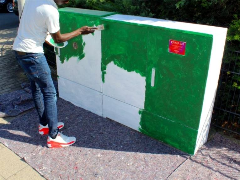 Auf dem Bild ist ein Stromverteilerkasten zu sehen, der zunächst weiß grundiert wurde. Eine Person ist in diesem Moment dabei diesen mit grüner Farbe zu streichen/bemalen.