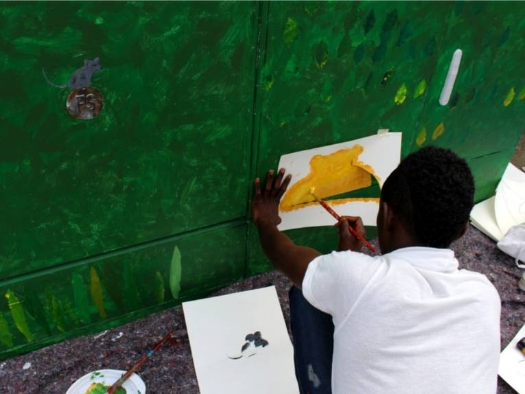 Auf dem Bild malt ein Mann etwas Gelbes auf dem Stromverteilerkasten, der darauf schon grün bemalt wurde.