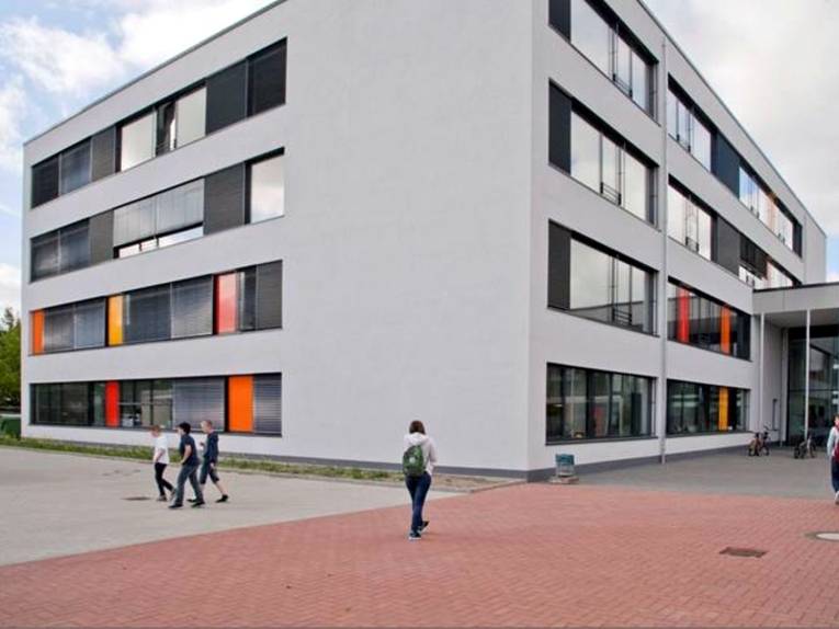 Auf dem Bild ist das moderne Gebäude der IGS Mühlenberg zu sehen. Ein paar Schüler sind vor der Schule zusehen.