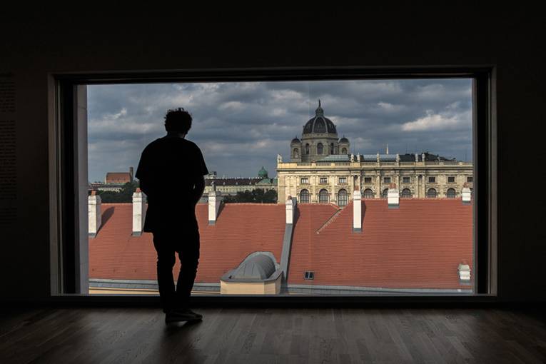 von innen nach außen fotografiert - ein Mann steht an einem großen Panoramafenster und schaut auf die Silouette eienr Stadt