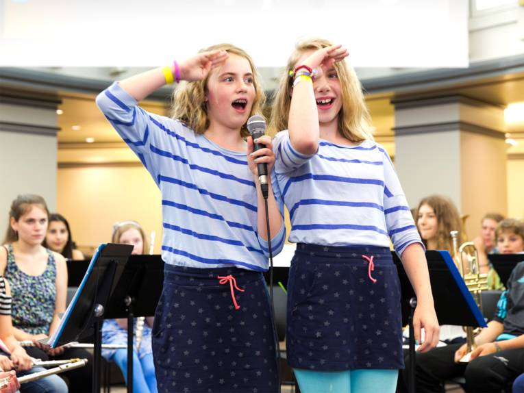 Anmoderation der Bläserklasse IGS Roderbruch: Zwei Schülerinnen performen vor dem versammelten Orchester auf der Bühne