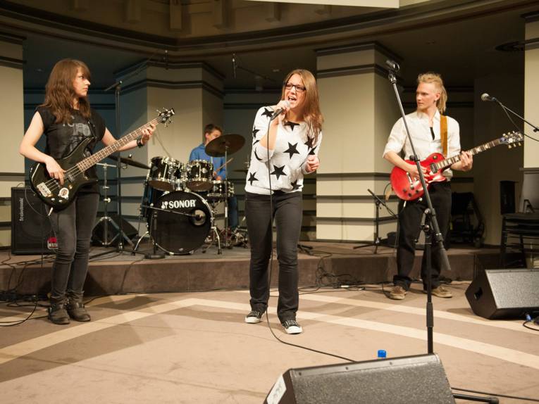 Eine Gitarristin, eine Sängerin und ein Gitarrist im Vordergrund der Bühne, im Hintergrund ein Schlagzeuger