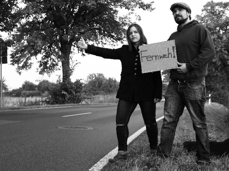 Eine Frau und ein Mann an einer Straße. Sie halten ein Schild, auf dem "Fernweh" steht. 