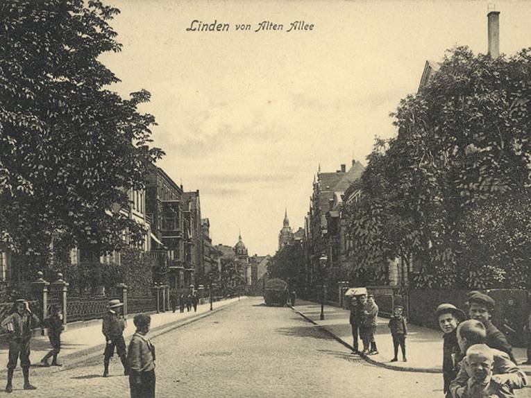 Die Postkarte ist betitelt "Linden von Alten-Allee" und zeigt den Blick in eine beidseitig mit zwei- bis dreigeschossigen Häuseren bestandene, relativ breite Straße. Im Vordergrund zahlreiche Kinder (Schüler), die den Fotografen bei der Arbeit beobachteten. Foto-Postkarte, um 1905