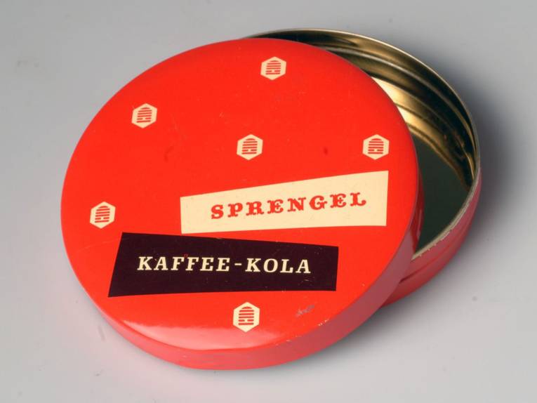 Flache runde Dose aus Blech mit abnehmbarem Deckel. Rot lackiert, auf der Oberseite fünf Bienenkörbe (Firmenzeichen Sprengel in der Form ab 1952) sowie in zwei annährend trapezförmigen Feldern die Aufschrift "Sprengel" und "Kaffee-Kola