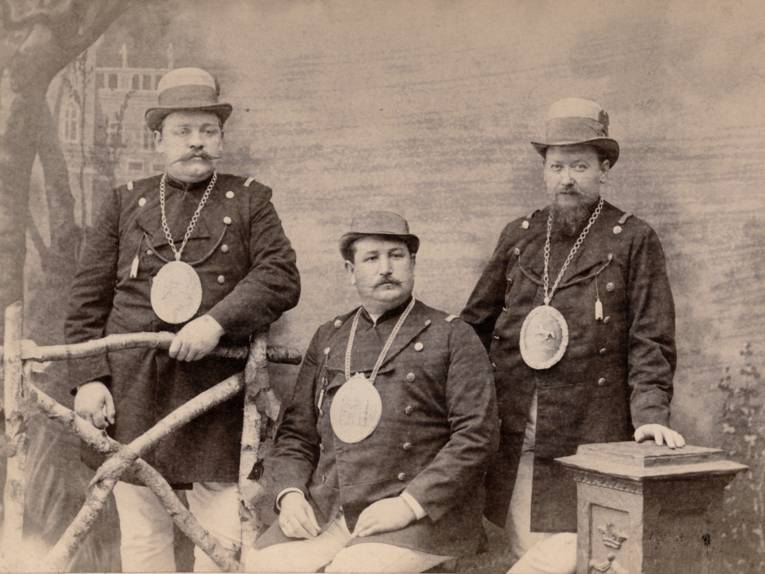 Atelieraufnahme: drei Männer in Schützenuniform. Sie tragen auffällige ovale Schilder an Halsketten auf der Brust. Das Schild des Mannes in der Mitte wohl mit dem hannoverschen Stadtwappen, der Mann rechts trägt Schild mit aufgelegtem Niedersachsenross. Nach der Uniform, die u.a. gekennzeichnet ist durch weiße Hosen, könnte es sich um Mitglieder des Uniformierten Schützenkorps handeln. Alle drei tragen ein Brustschild, das sie als "Beste Mann" kennzeichnet, also als Gewinner eines Schießwettbewerbes beim hannoverschen Schützenfest.