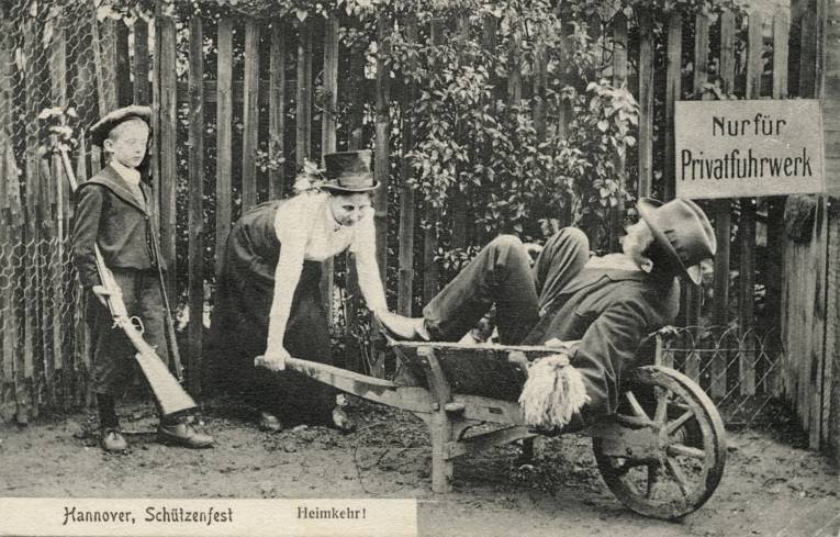 Foto-Postkarte, Poststempel 1910: Lichtdruck nach S/W-Foto. Betitelt "Hannover, Schützenfest" und "Heimkehr!". Das Bild zeigt einen Mann auf einer Schubkarre liegend, an deren Holmen eine Frau posiert, als würde sie die Karre gebückt schieben. Links daneben ein Junge, der das Gewehr des Vaters über seine Schulter gehängt hat. Im Hintergrund ein Stakettenzaun mit Schild "Nur für Privatfuhrwerk", das offensichtlich den Scherz mit dem Betrunkenen noch verstärken soll.
