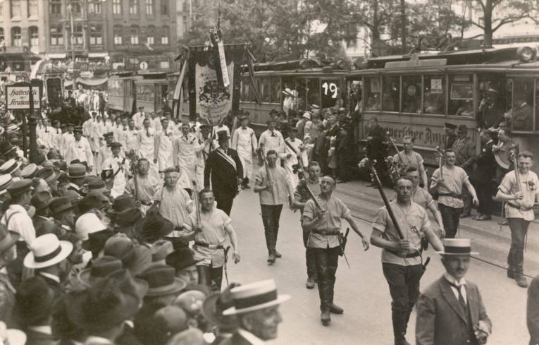 Foto-Postkarte, o. J. (1920-1929): Die Postkarte zeigt den Schützenausmarsch in der Georgstraße. Zentrales Bildmotiv ist die Gruppe der hannoverschen Schlachter. Zahlreiche Zuschauer am Straßenrand und in den angehaltenen Straßenbahnen betrachten das Geschehen.