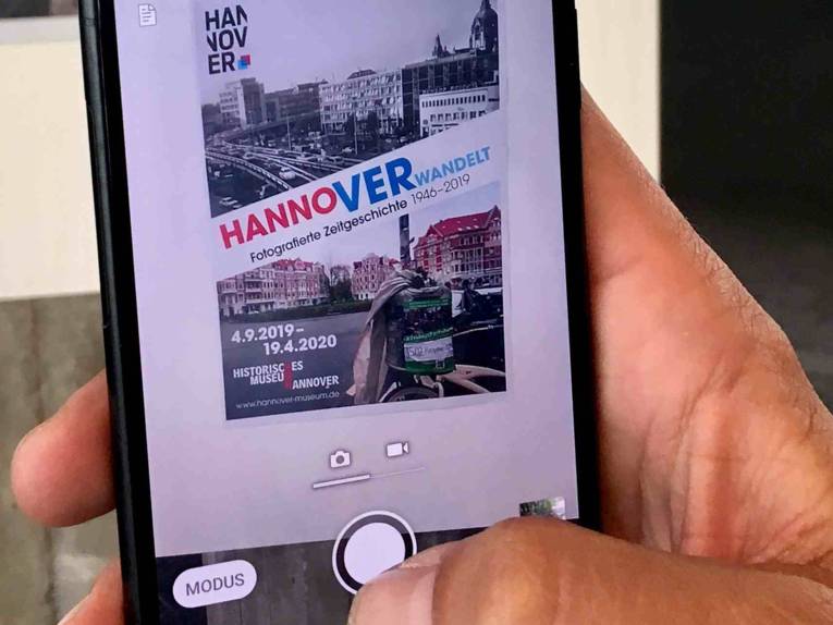 Hand mit Smartphone und Teil vom Ausstellungsplakat zur Sonderausstellung "Hannoverwandelt"