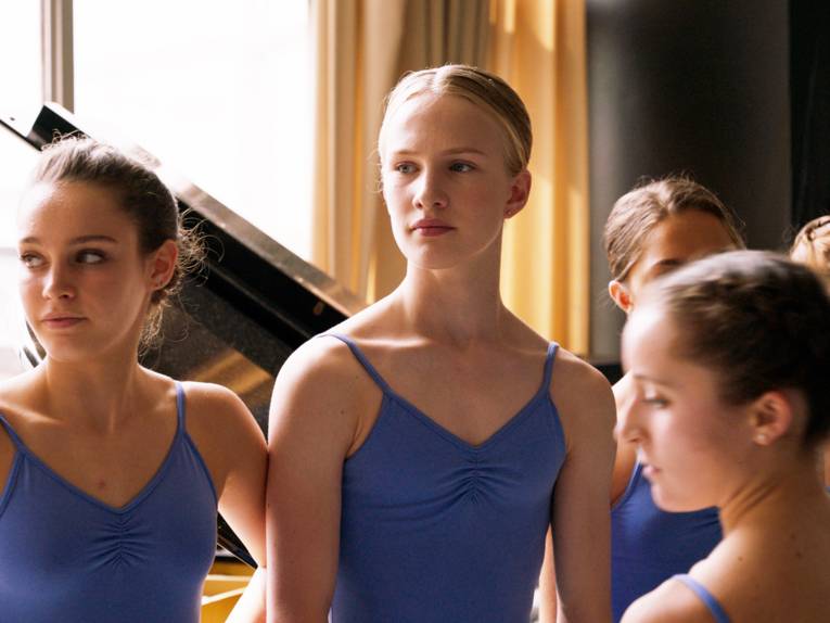 Im Bild zu sehen, ist eine Gruppe von Ballerinas, die an einem Flügel stehen. In der Mitte die transsexuelle Protagonistin.