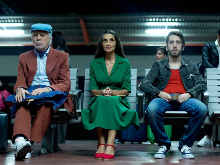 Im Bild zu sehen sind drei Personen, ein alter Mann, eine ältere Frau und ein junger Mann, die in einem Flughafen warten.