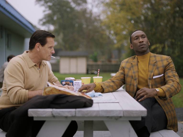 Im Bild zu sehen sind Viggo Mortensen und Mahershala Ali, die an einem weißen Tisch sitzen und sich bei Getränken und Essen unterhalten.