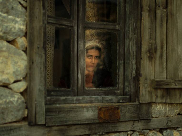 Im Bild zu sehen ist eine junge Frau, die aus einem schmutzigen Fenster schaut.